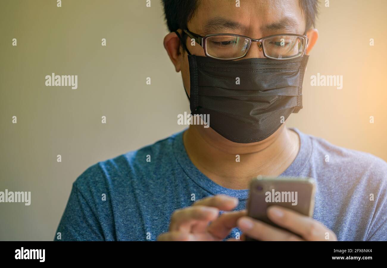 Porträt Asian Mann trägt schwarze medizinische Maske und SMS auf Smartphone, Nahaufnahme Asian Mann mit Brille, schwarze kurze Haare, Augen zu smartphon schauen Stockfoto