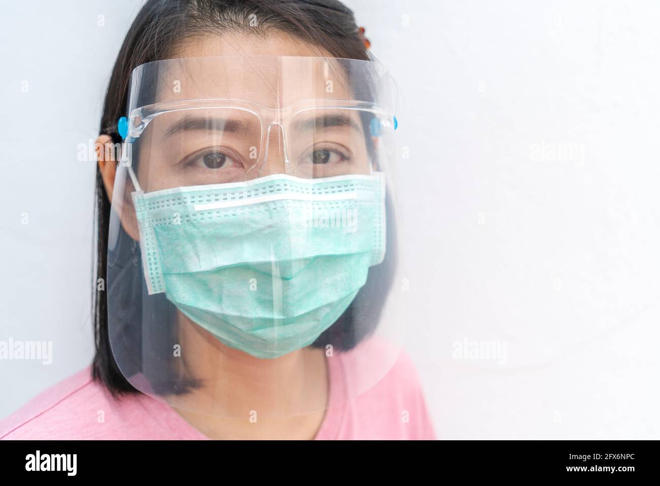Nahaufnahme Gesicht Asiatische Frau mittleren Alters trägt medizinische Gesichtsmaske und Kunststoff-Gesichtsschutz, schwarze kurze Haare, Blick auf die Kamera, Modell auf weißer Wand BA Stockfoto