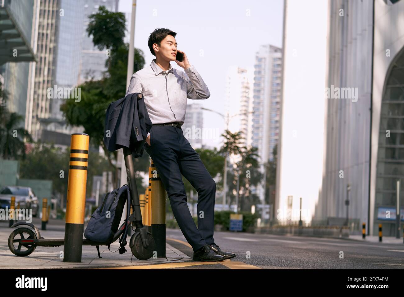 Junger asiatischer Geschäftsmann steht neben seinem Elektroroller auf Straße, die einen Anruf mit Handy im Schlepptau der modernen Stadt Stockfoto