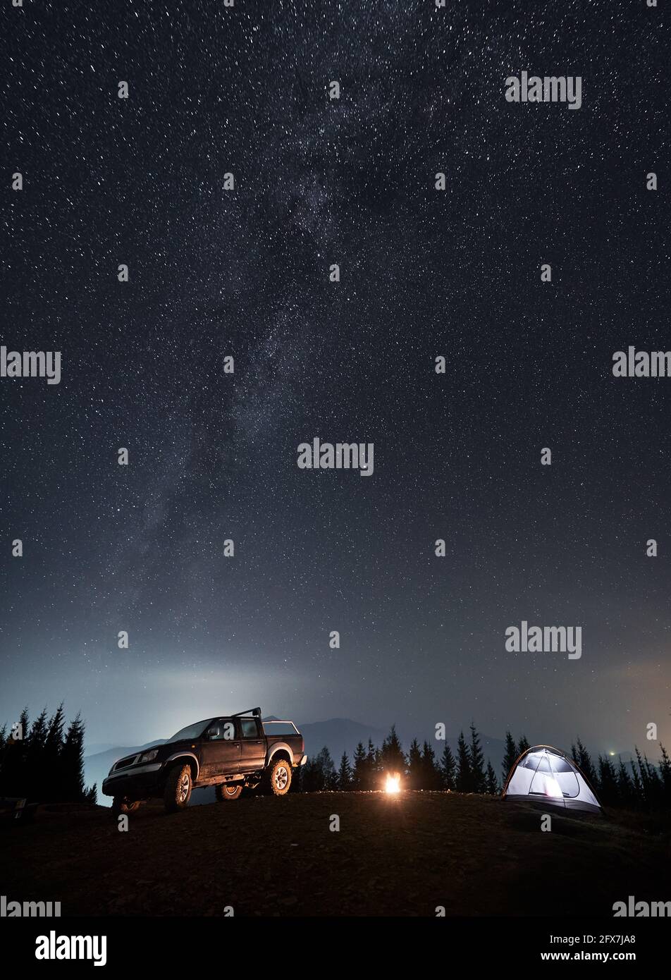 Farbenprächtiger nächtlicher Sternenhimmel und Milchstraße über dem Ferienlager auf einem Berghang. Schwarzes SUV, beleuchtetes Zelt und Lagerfeuer zwischen ihnen auf dem Hintergrund von Silhouetten von Bäumen und Bergen. Stockfoto