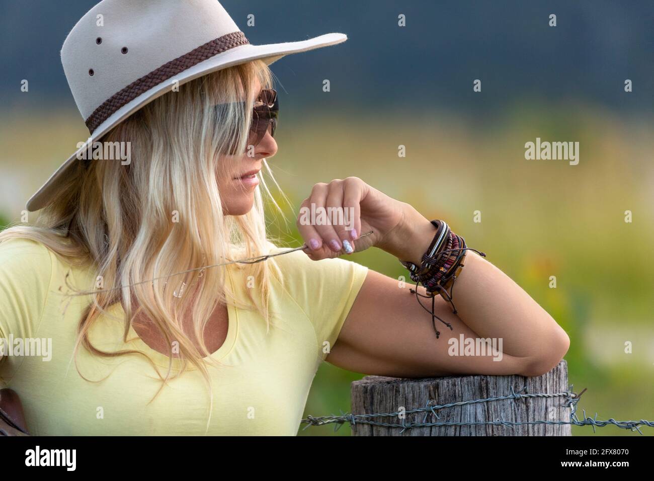 Gesunde Frau auf einem Bauernhof trägt einen Bauernhof Hut und Leder geflochtene Armbänder lehnt sich an einen Zaunpfosten mit einem Eine Grassträhne Stockfoto