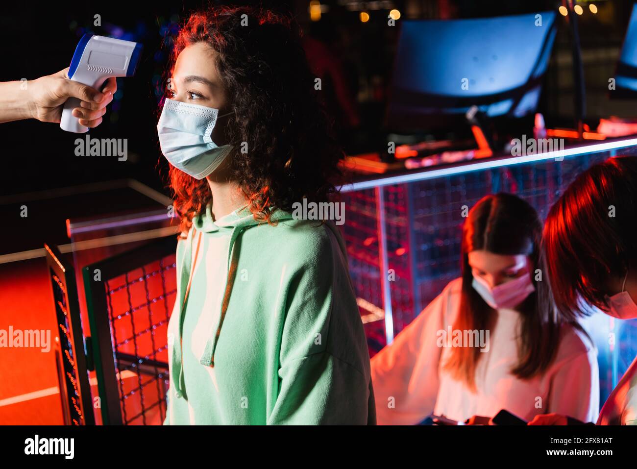 afroamerikanisches Mädchen in medizinischer Maske in der Nähe des Kontrollers mit Pyrometer Und Teenager-Freunde Stockfoto