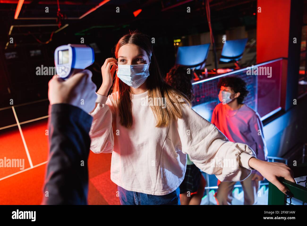 Teenager-Mädchen in medizinische Maske in der Nähe Controller mit Pyrometer und Freunde auf unscharfem Hintergrund Stockfoto