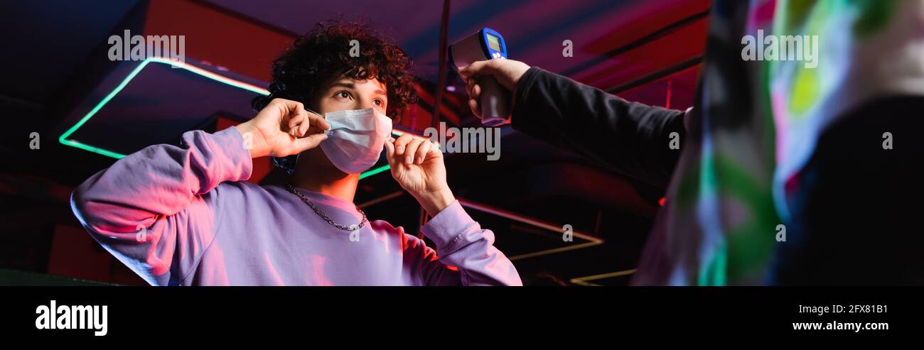 Teenager-Gamer, der eine medizinische Maske in der Nähe des Controllers mit Pyrometer, Banner aufsetzt Stockfoto