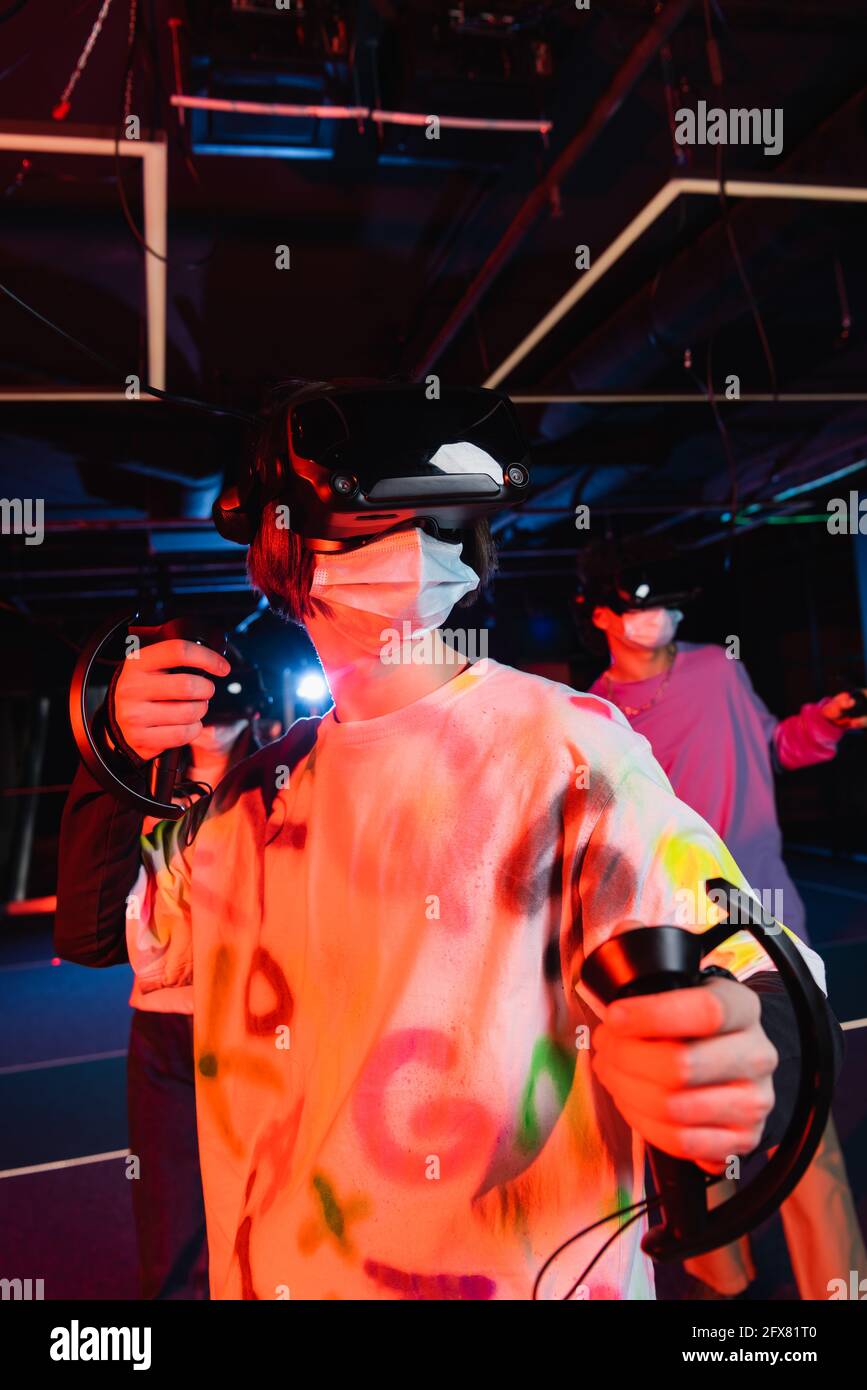 Teenager in medizinischen Maske Gaming in der Nähe verschwommen Freunde in vr-Wiedergabezone Stockfoto