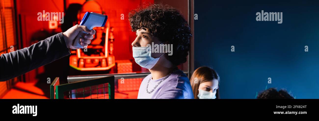 Teenager Junge in medizinischer Maske in der Nähe Controller mit Pyrometer und Mädchen auf verschwommenem Hintergrund, Banner Stockfoto
