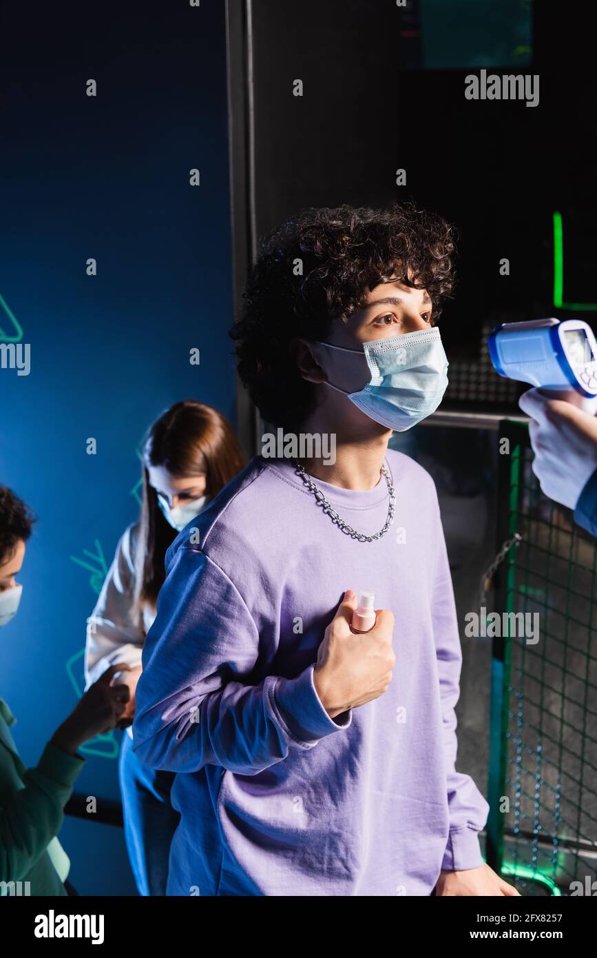 Teenager in medizinischer Maske, die Händedesinfektionsmittel in der Nähe des Kontrollers hält Und multiethnischen Freunden Stockfoto