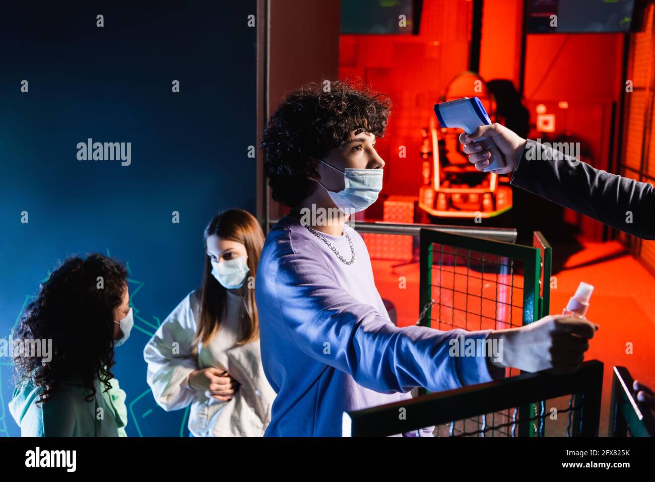 Teenager Gamer in medizinischer Maske hält Hand Desinfektionsmittel in der Nähe des Controllers Mit Pyrometer und interracial Freunden Stockfoto