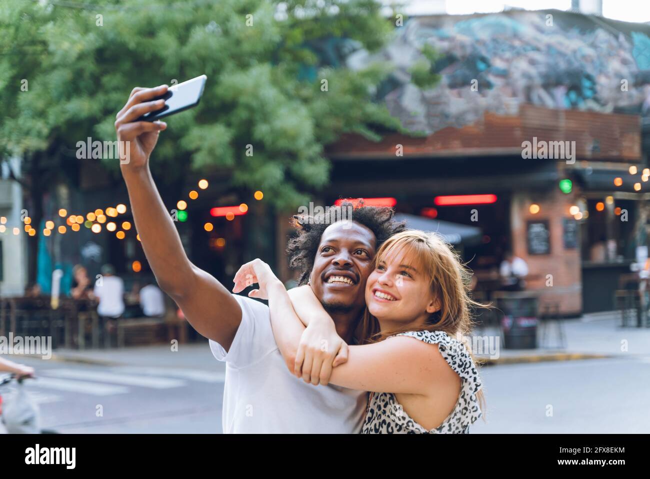 Glückliches junges multiethnisches Paar, das abends Selfies auf der Straße macht. Stockfoto