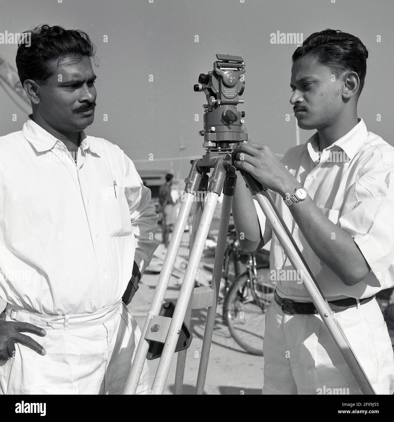 1957, historisch, zwei indische Vermesser mit einem No 1 Hilger & Watts Microptic Theodolit auf Stativ auf der Baustelle des Durgapur Steel Project, Durgapur, Westbengalen, Indien. Als präzises optisches Instrument zur Messung von Winkeln zwischen sichtbaren Punkten in der horizontalen und vertikalen Ebene wurde dieser Transittheodolit im Bau- und Infrastrukturbau ausgiebig eingesetzt. Stockfoto