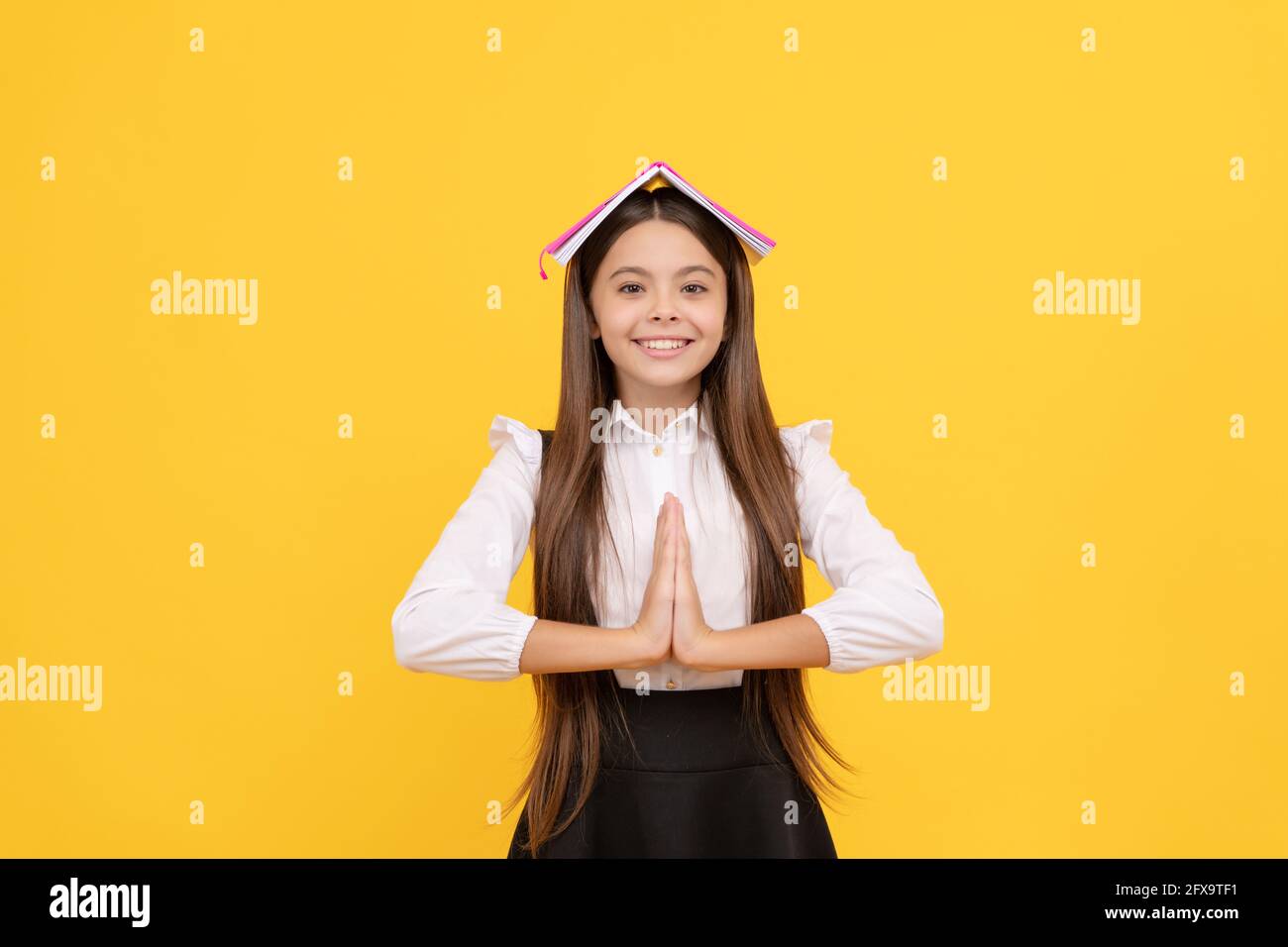 Glückliches Teenager-Mädchen in Schuluniform mit Buch auf Kopf meditieren mit Handbewegung, Ruhe bewahren Stockfoto