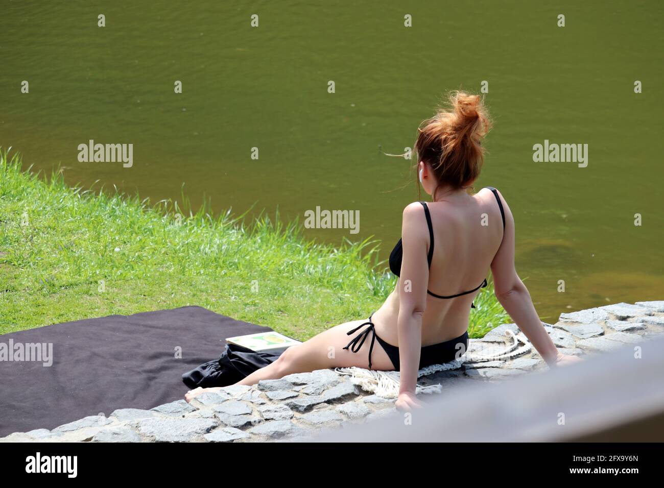 Frau in schwarzem Bikini-Sonnenbaden sitzt am Strand. Entspannen und Urlaub am Sommersee Stockfoto
