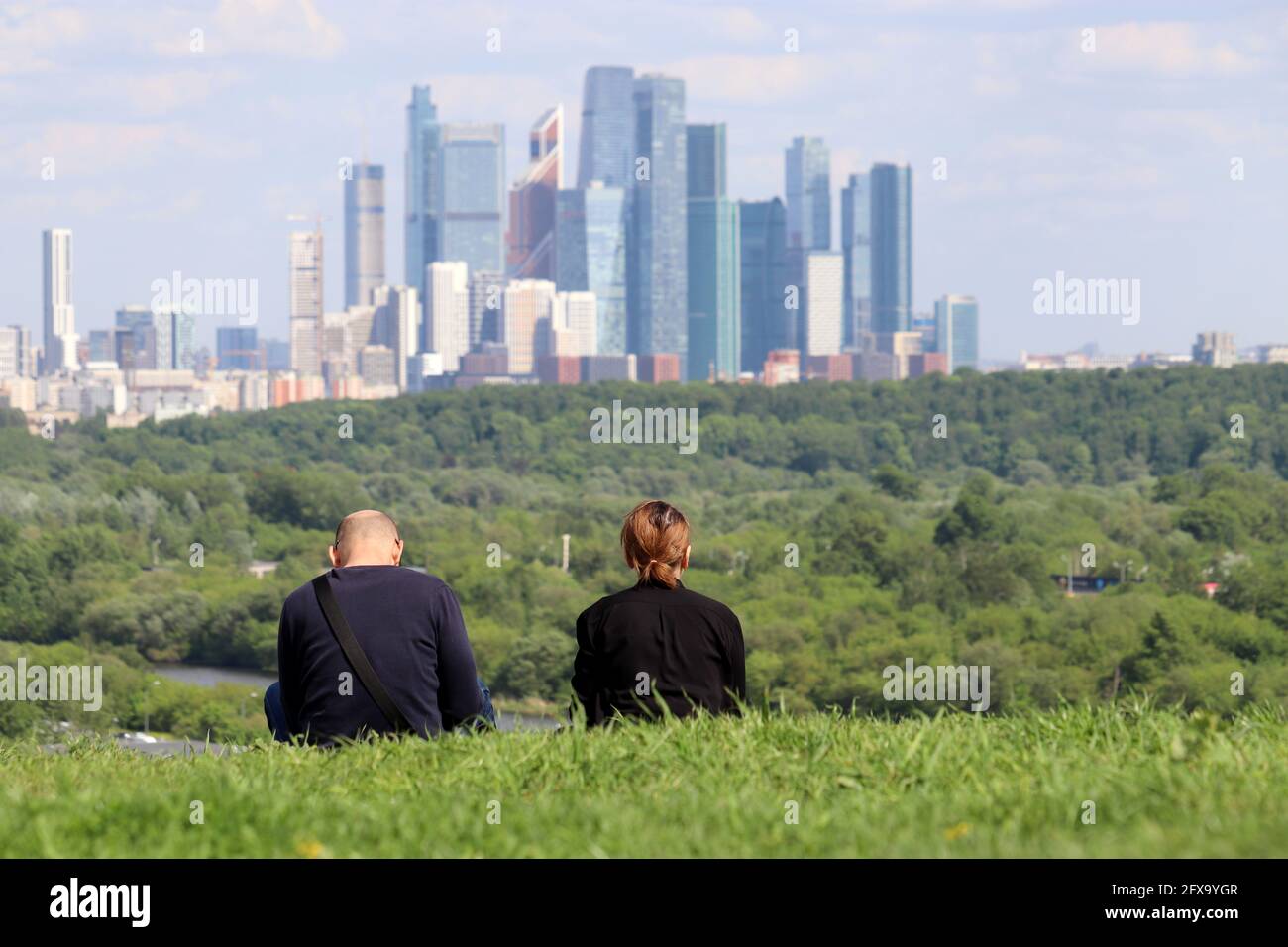 Ehepaar sitzt im Gras gegen die Wolkenkratzer der Stadt Moskau. Futuristisches Stadtbild, Konzept der suburbanen Sommerfreizeitgestaltung Stockfoto