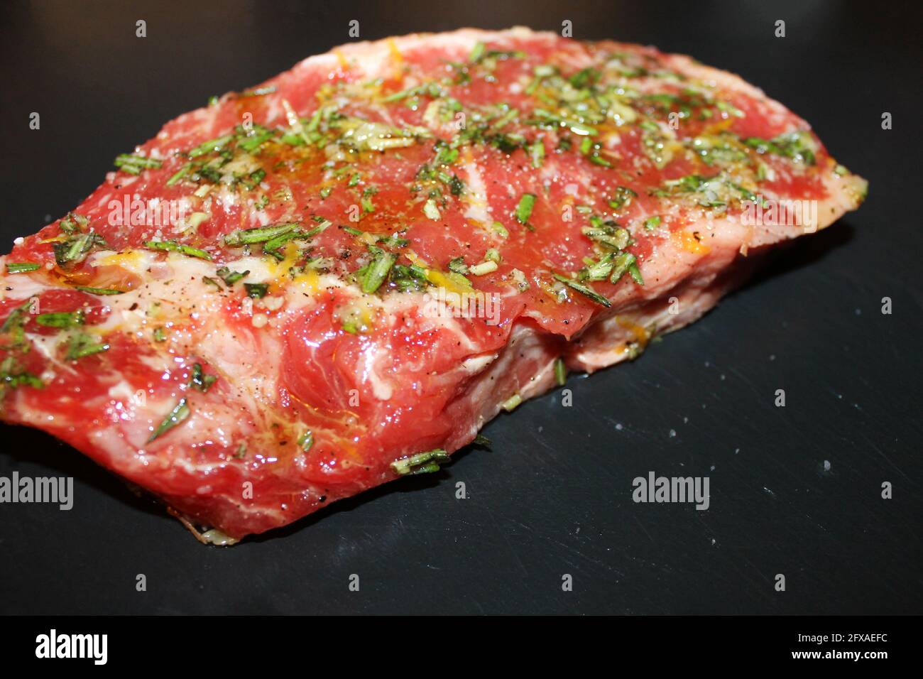 Nahaufnahme von ungekochten Rindfleisch, gewürzt mit Salz, Pfeffer, Basilikum und Zitronenschale, auf einem Schneidebrett aus schwarzem Stein. Stockfoto