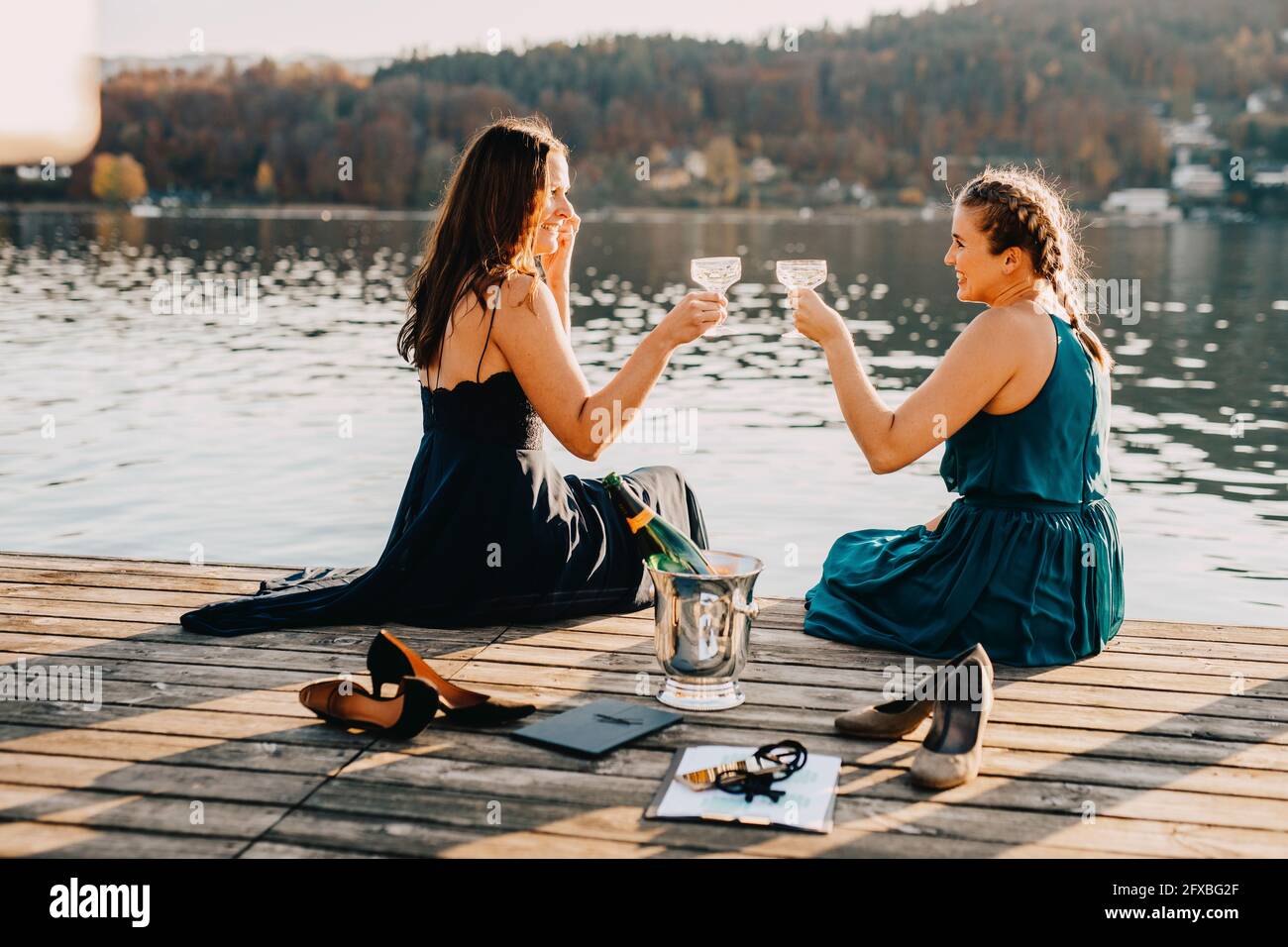 Weibliche Veranstaltungsplanerinnen toasten Champagner, während sie auf dem Steg über dem See sitzen Stockfoto
