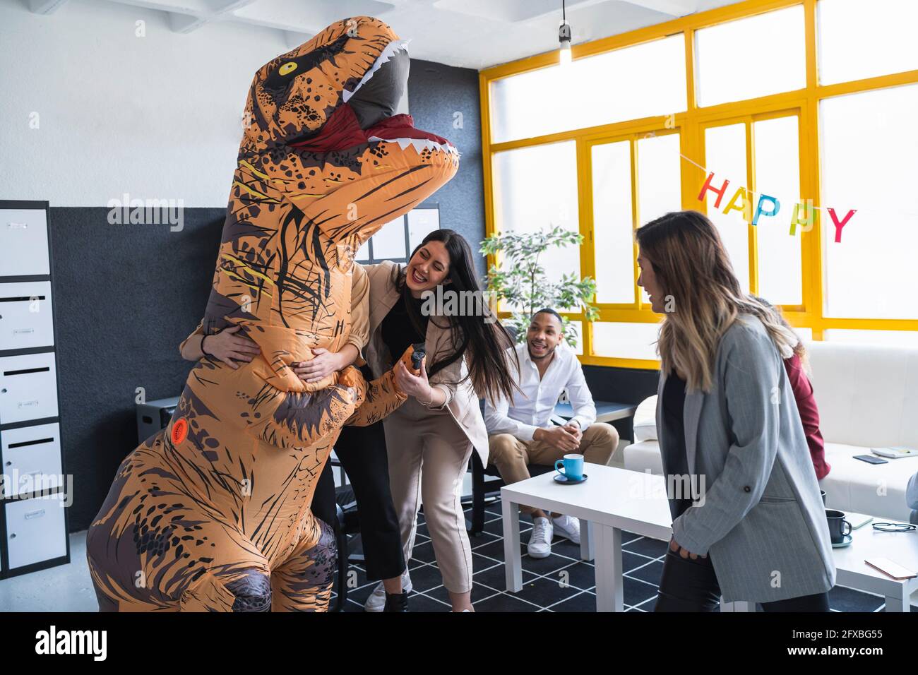 Glückliche Geschäftsfrauen umarmen weibliche Kollegin in Dinosauriers Kostüm während der Geburtstagsfeier im Büro Stockfoto