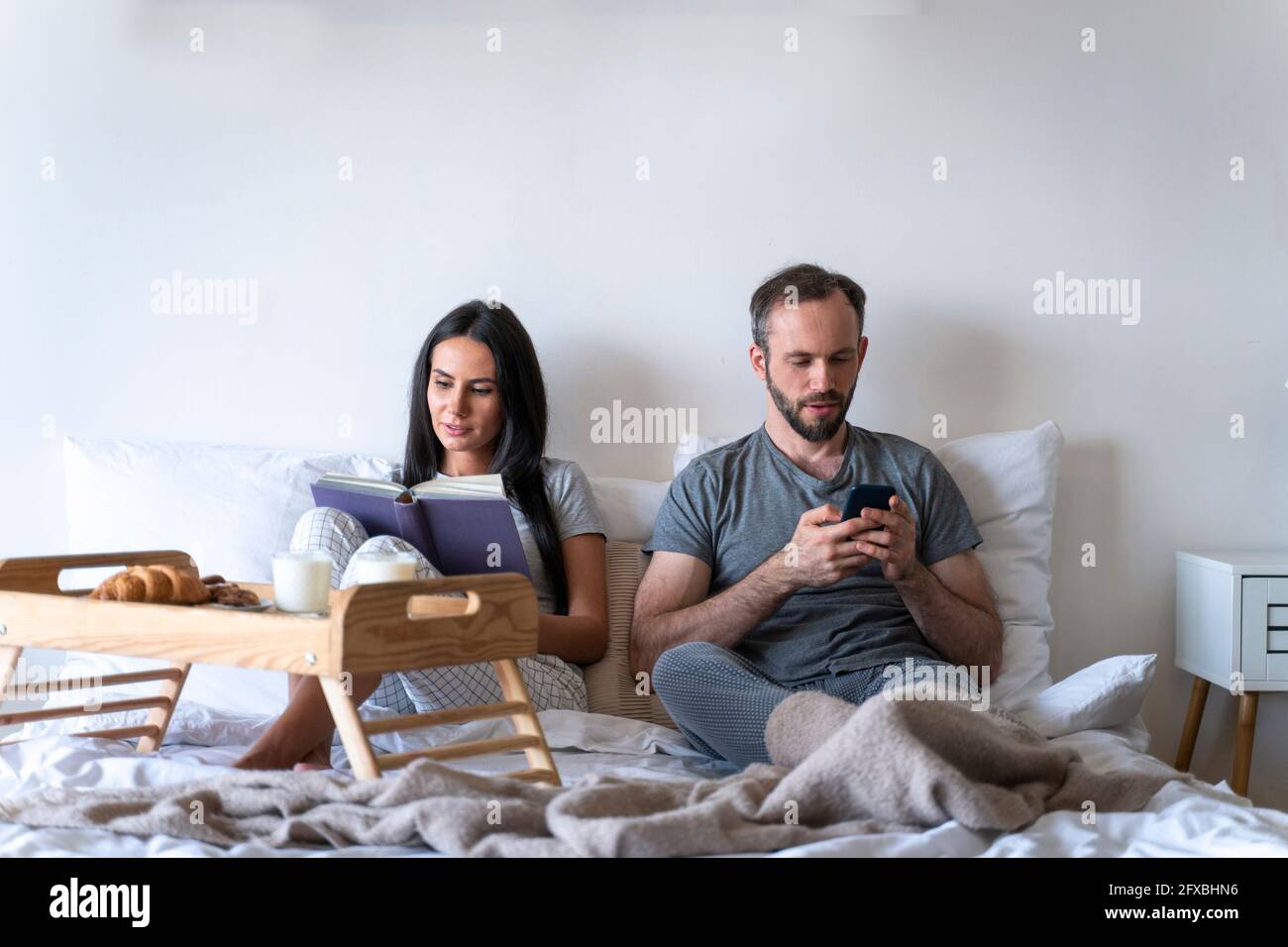 Eine Frau, die mit einem Mann ein Buch liest, sitzt mit einem Smartphone, während sie zu Hause auf dem Bett frühstückt Stockfoto