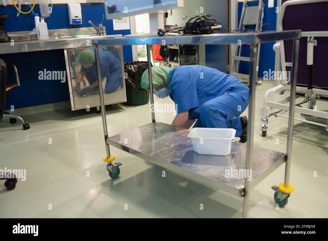 Frau vom Reinigungsservice eines Krankenhauses hockend, die einen tragbaren Operationstisch im vorherigen Operationssaal putzt. Sanitärkonzept Stockfoto