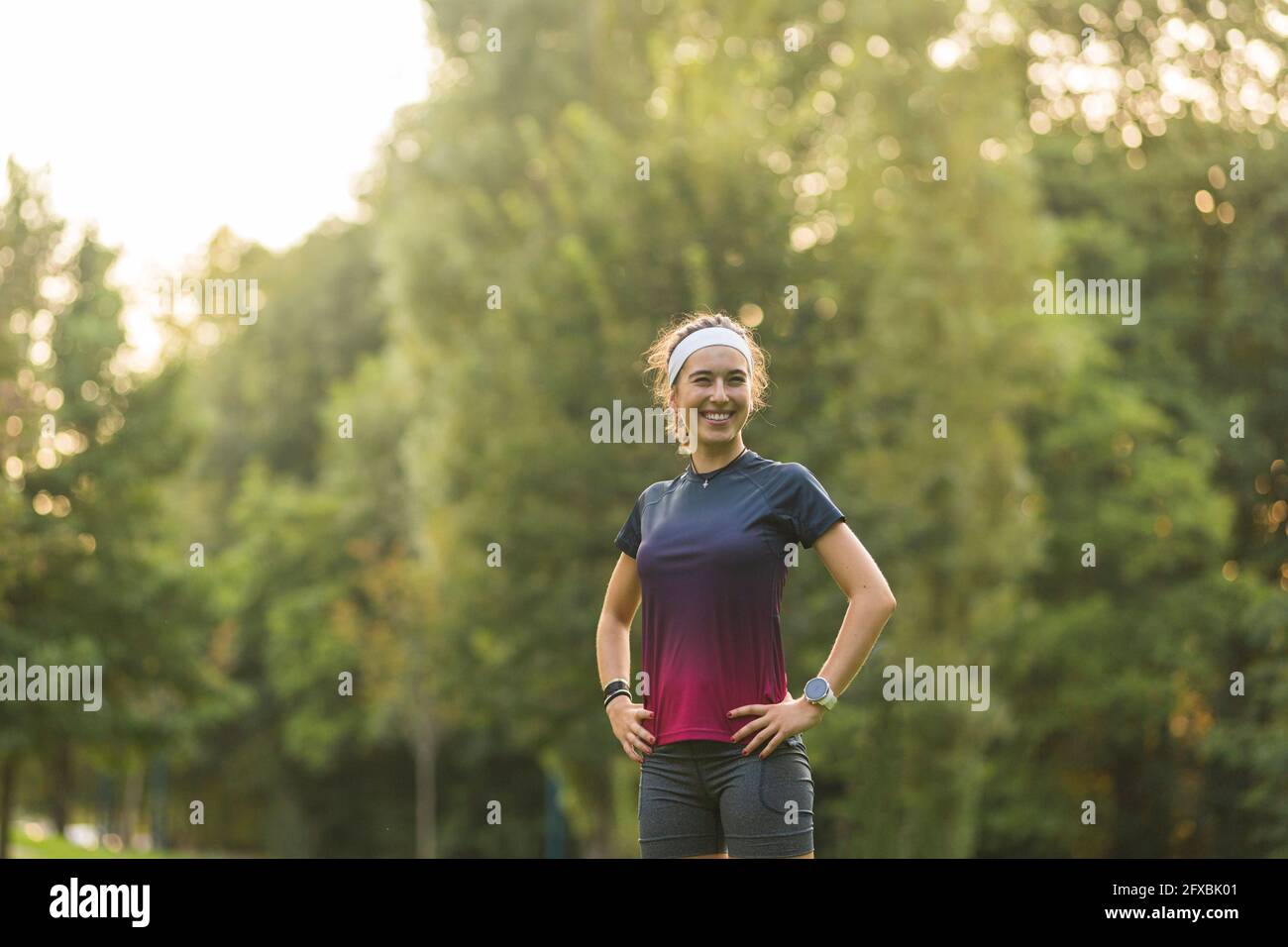 Lächelnde Frau mit den Händen auf der Hüfte, die im öffentlichen Park steht Stockfoto