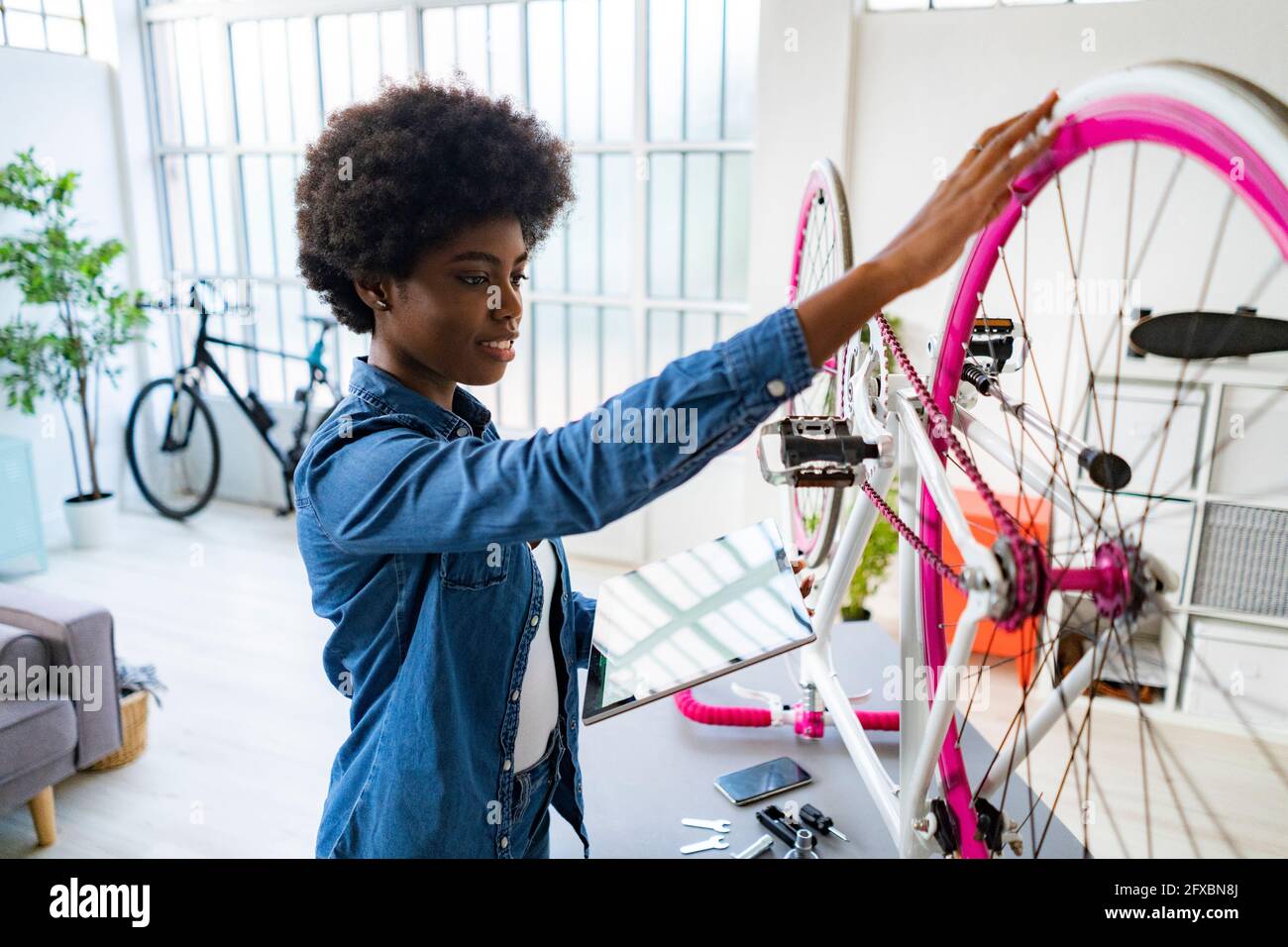 Lächelnde Frau repariert das Fahrrad, während sie zu Hause ein digitales Tablet verwendet Stockfoto