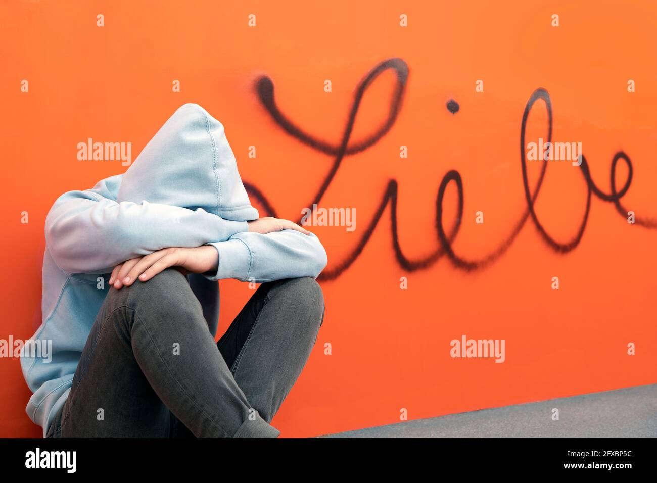 Trauriger Teenager, der vor dem deutschen Wort für Liebe an der orangen Wand sitzt Stockfoto