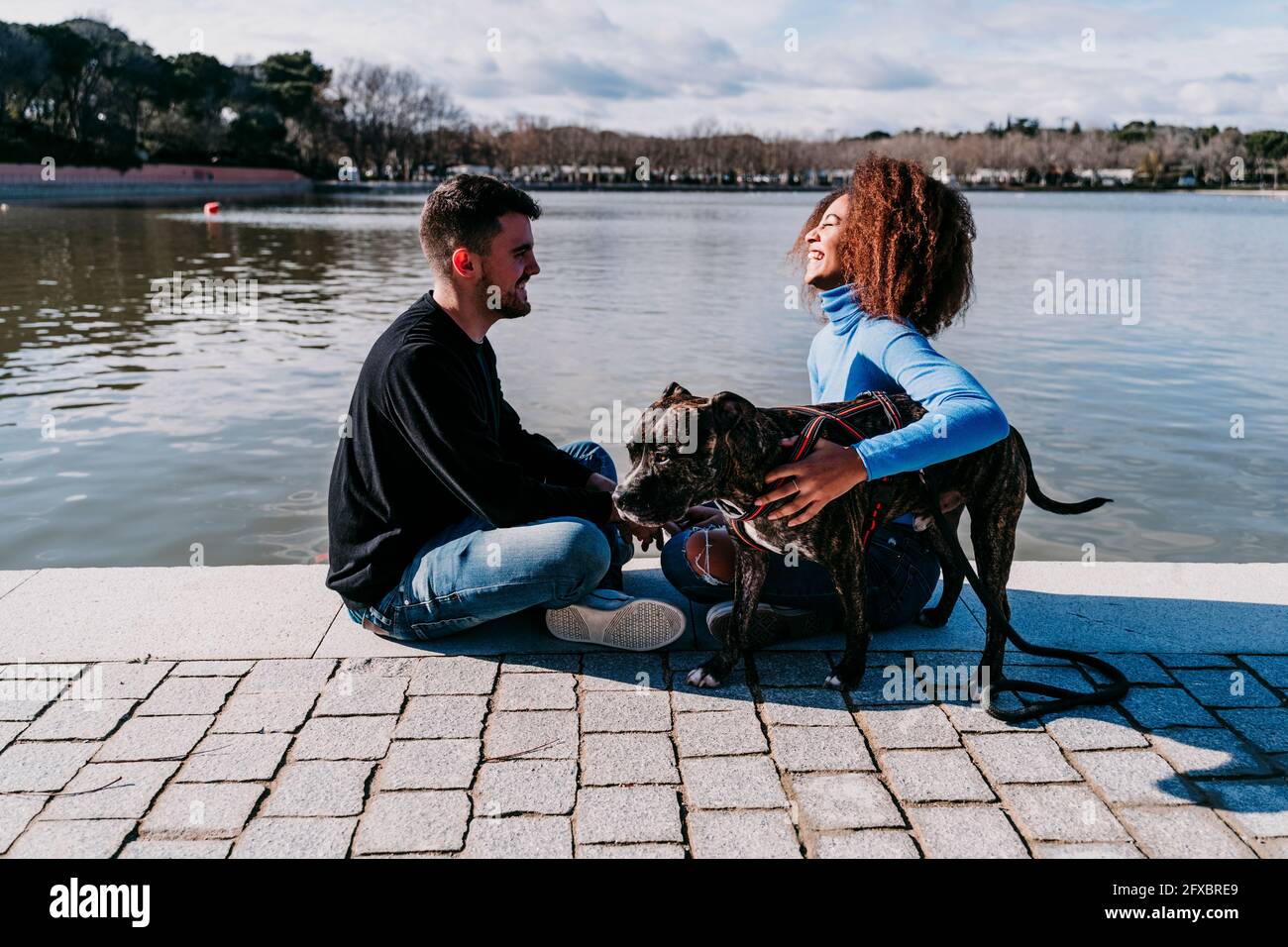 Lächelnde Freundin mit Freund, der beim Hund auf dem Fußweg am See sitzt Stockfoto