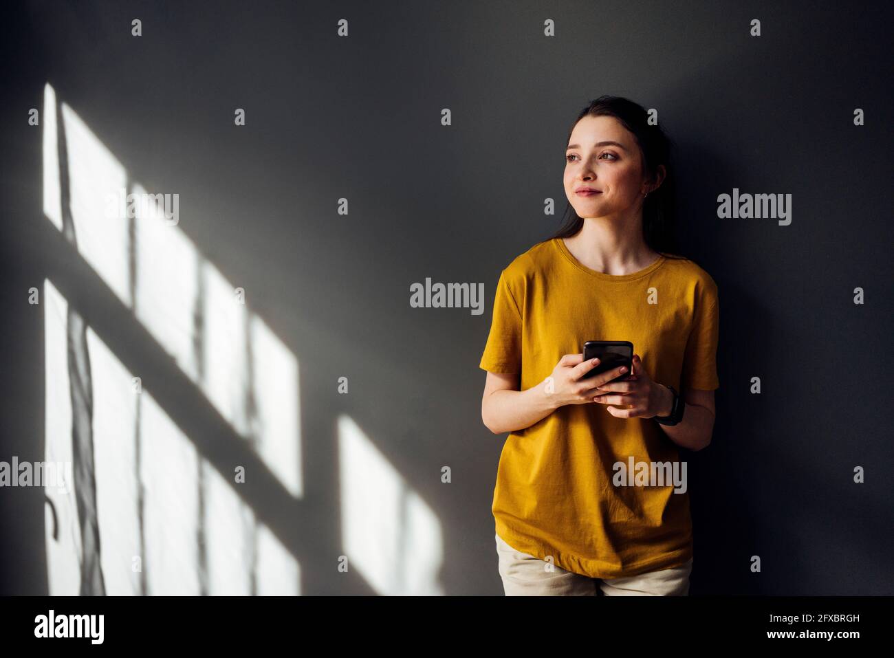 Nachdenkliche Frau mit Mobiltelefon, das am Fensterschatten an der Wand hängt Stockfoto