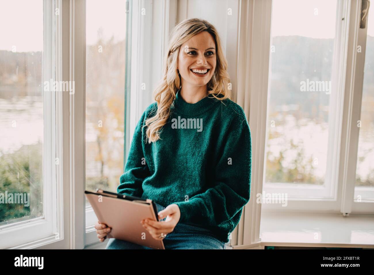 Blonde Frau im Sweatshirt sitzend mit Tablet am Fenster im Büro Stockfoto