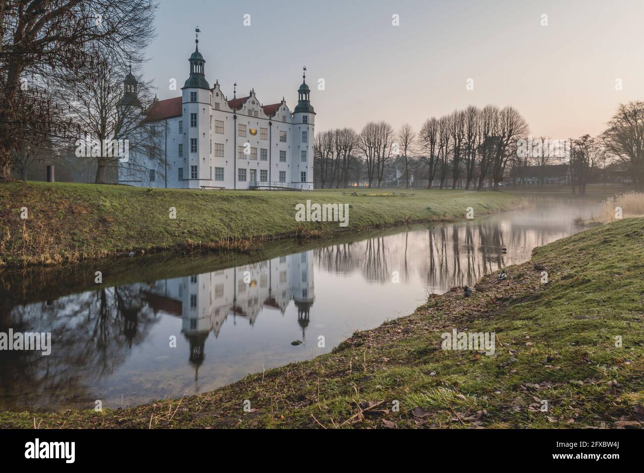 Deutschland, Schleswig-Holstein, Ahrensburg, Schloss Ahrensburg spiegeln sich im umliegenden Graben bei nebliger Morgendämmerung Stockfoto