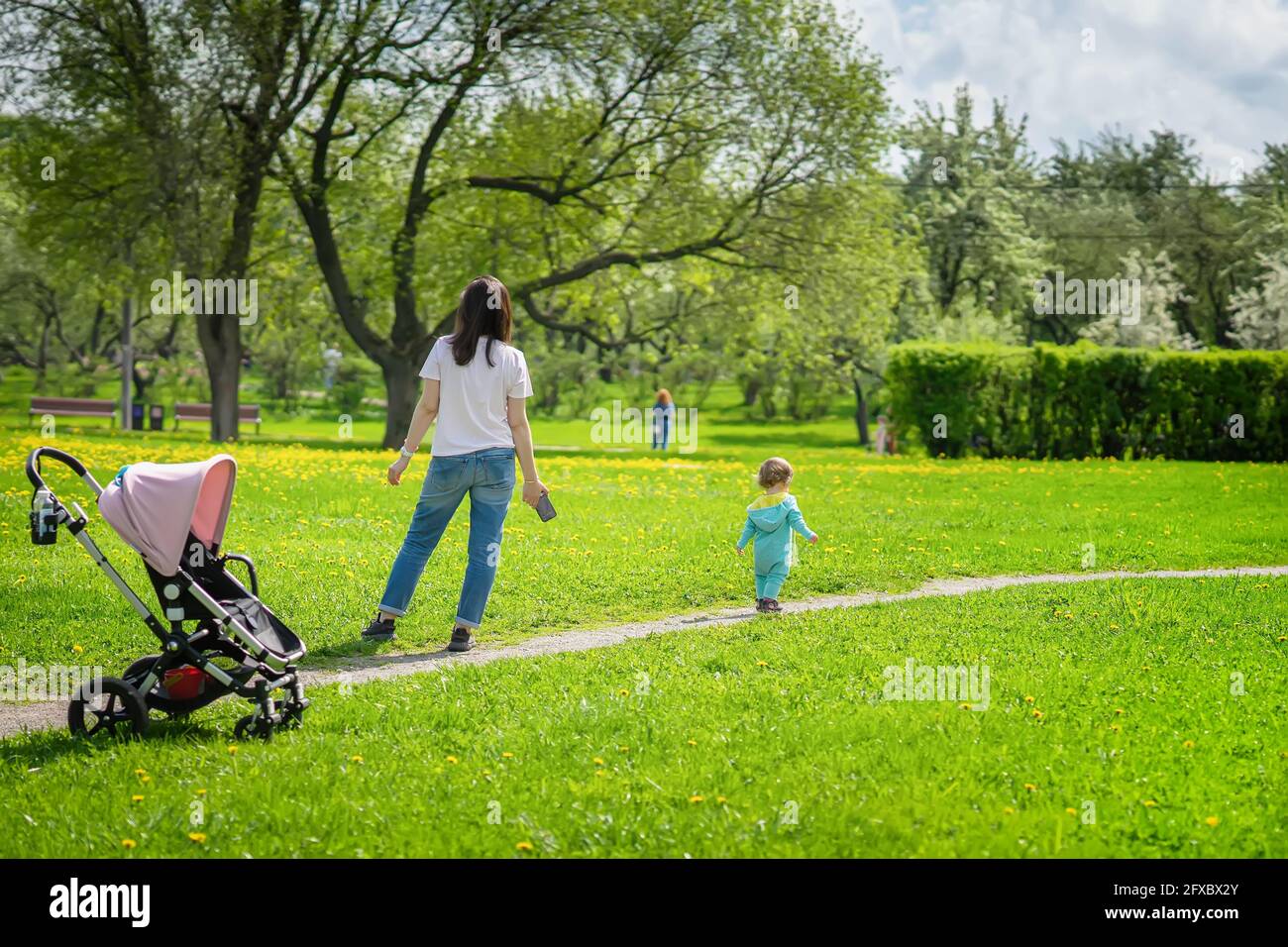 Junge Frau geht mit kleinem Baby im Park, helle Frühlingswiese mit Eselinnen, sonniger Tag Stockfoto