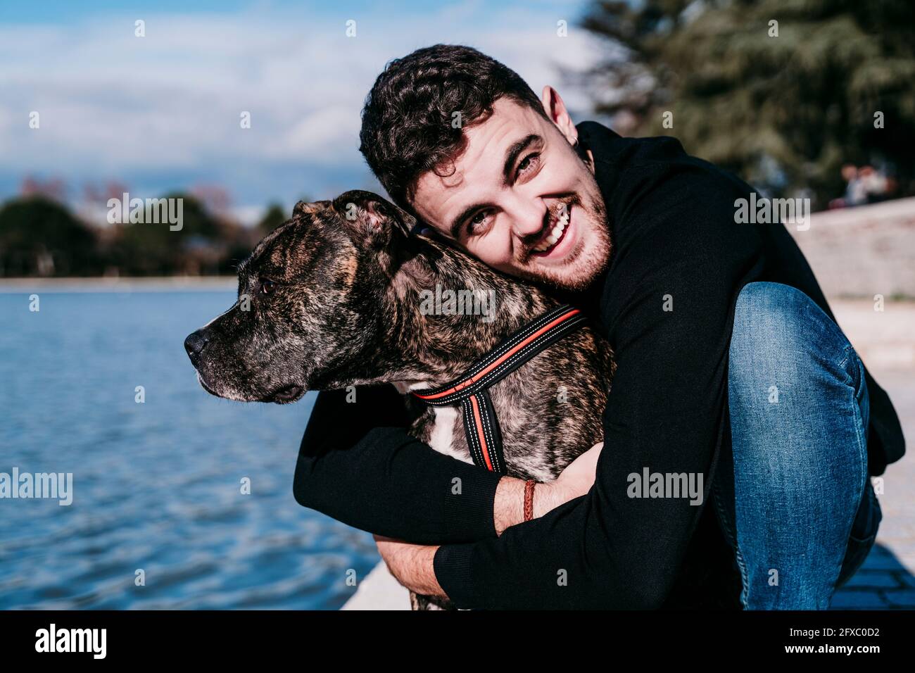 Lächelnder junger Mann umarmt Hund, während er am See hockend Stockfoto