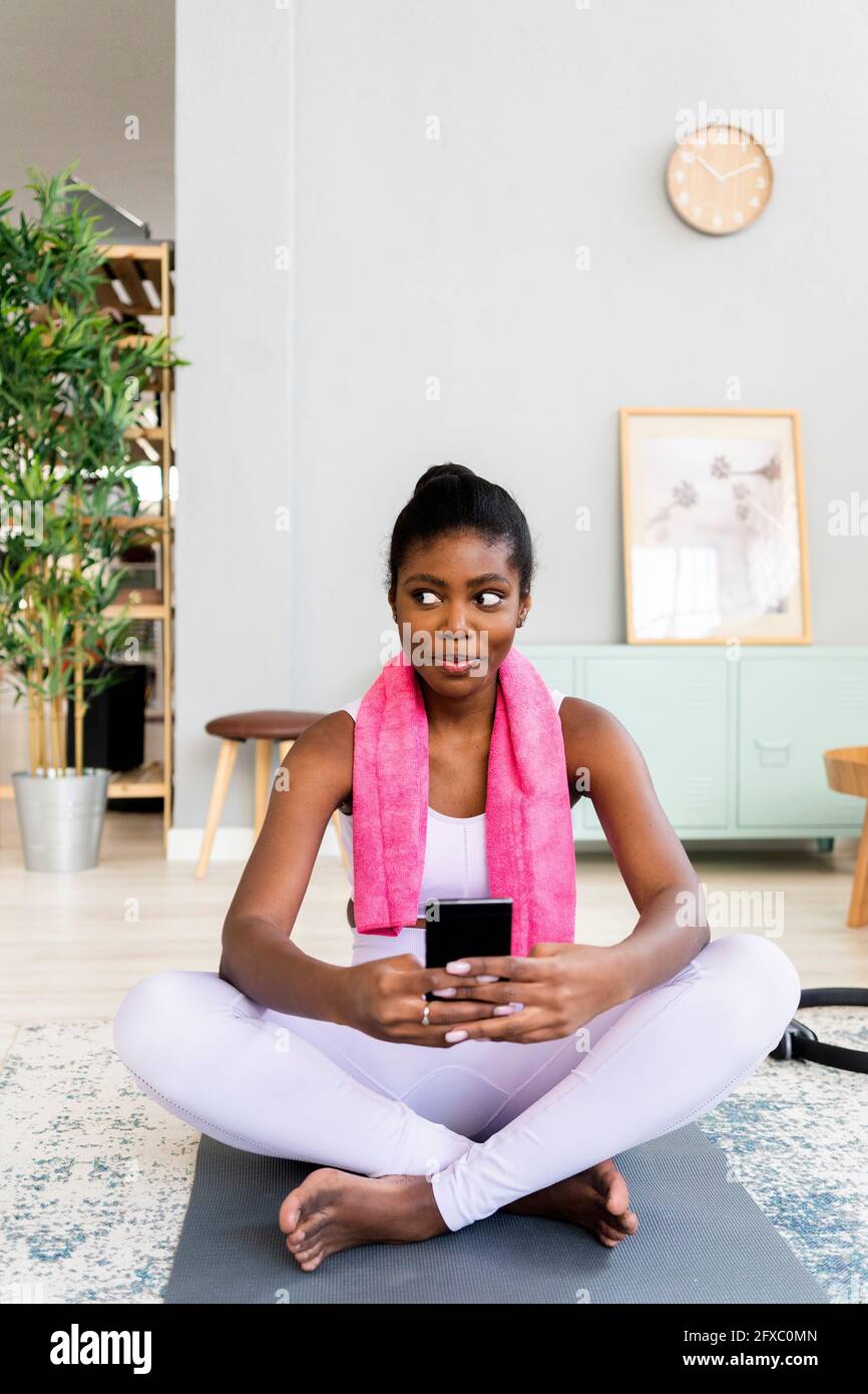 Junge Frau mit Handy, während sie auf der Trainingsmatte sitzt Zu Hause Stockfoto