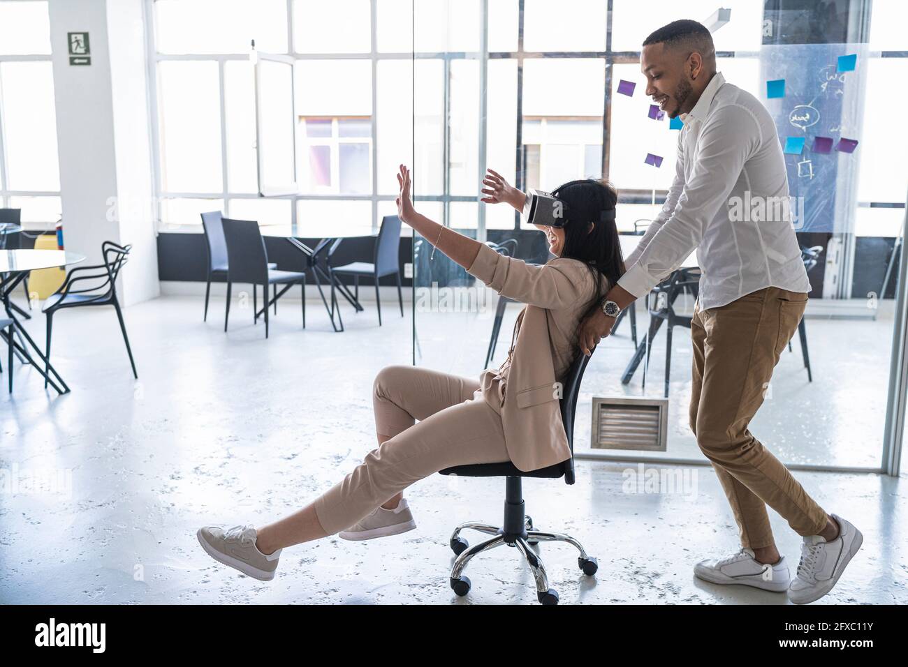 Männlicher Profi schiebt Geschäftsfrau trägt Virtual-Reality-Simulator auf Stuhl im Coworking-Büro Stockfoto