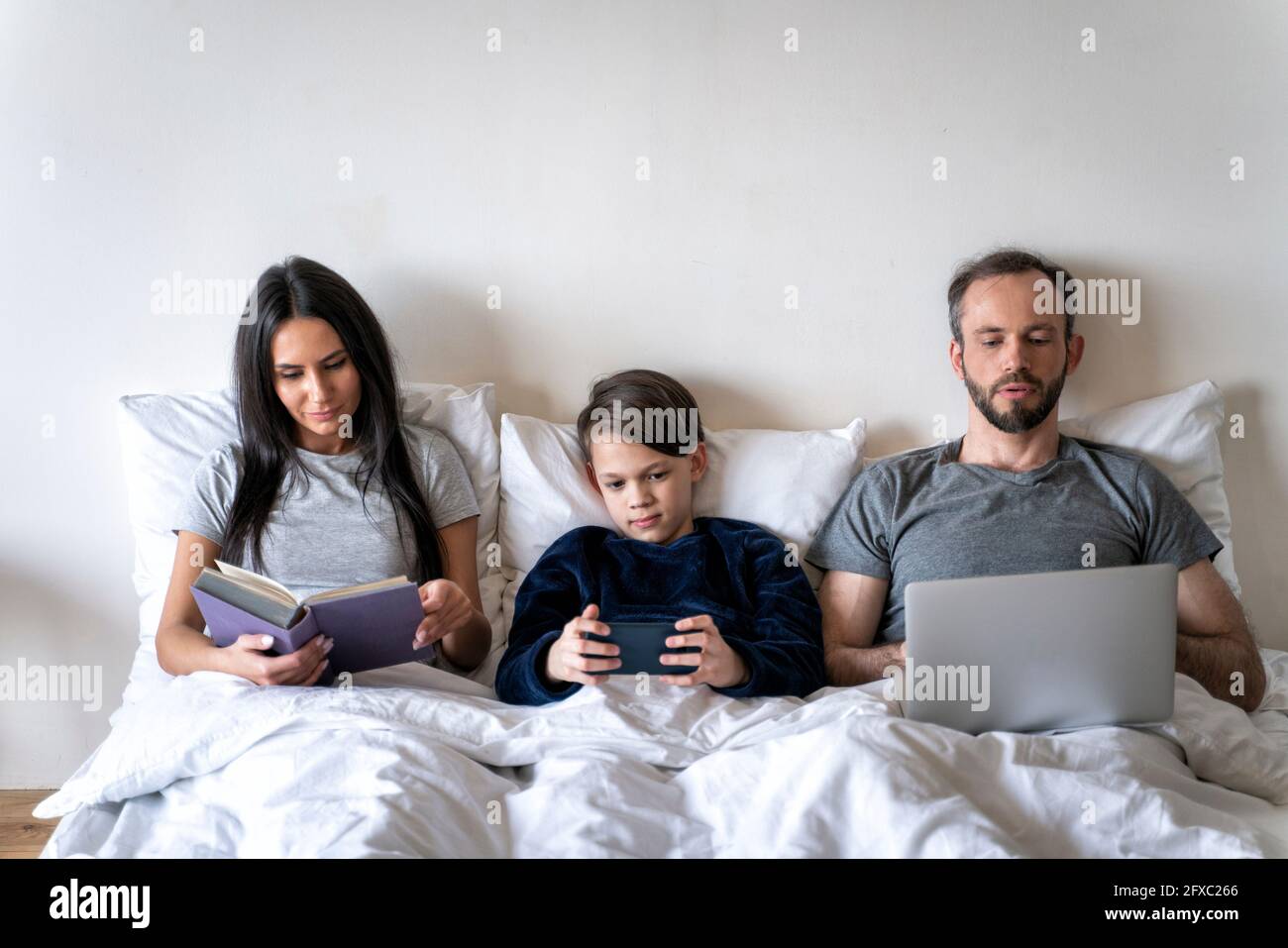 Frau liest Buch, während Vater und Sohn drahtlose Technologien im Bett zu Hause verwenden Stockfoto