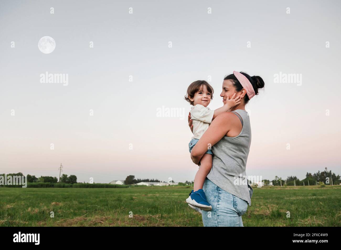 Mutter schaut, während sie den Sohn während des Sonnenuntergangs trägt Stockfoto