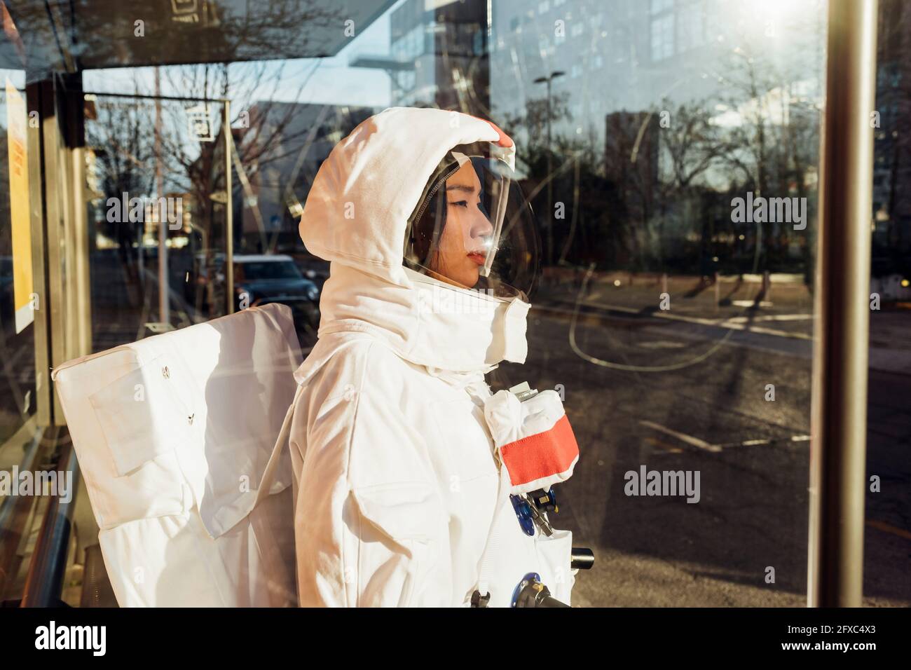 Weibliche Astronautin im Raumanzug, die an einem sonnigen Tag durch das Glas an der Bushaltestelle steht Stockfoto