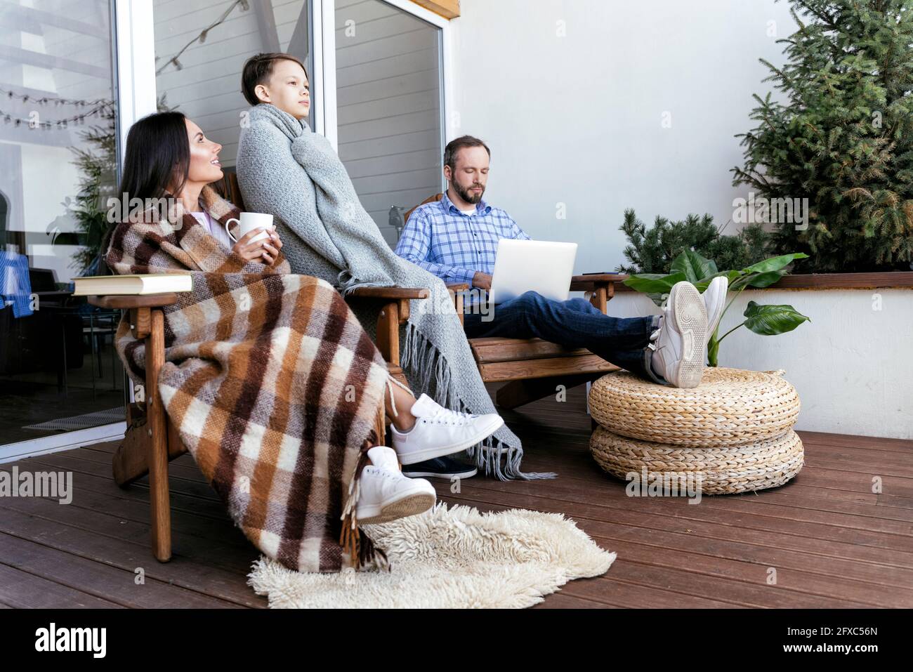 Mutter und Sohn wickelten sich in eine Decke, während sie mit dem Vater auf dem Balkon mit einem Laptop saßen Stockfoto
