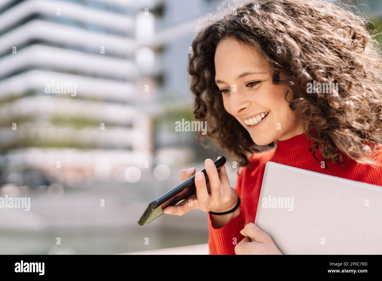 Lächelnde junge Frau, die an sonnigen Tagen mit einem Smartphone spricht und dabei den Laptop in der Hand hält Stockfoto