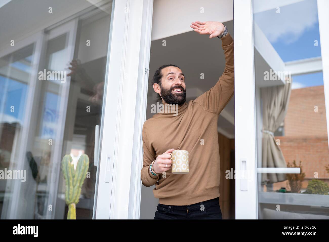 Glücklicher Mann winkt, während er zu Hause an der Schiebetür steht Stockfoto