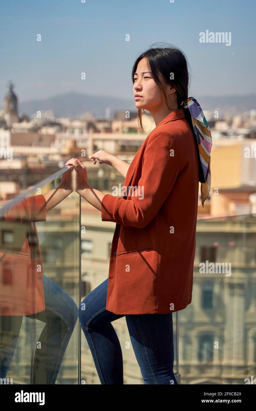 Junge Frau, die auf der Dachterrasse am Glasgeländer steht und die Aussicht ansieht Stockfoto