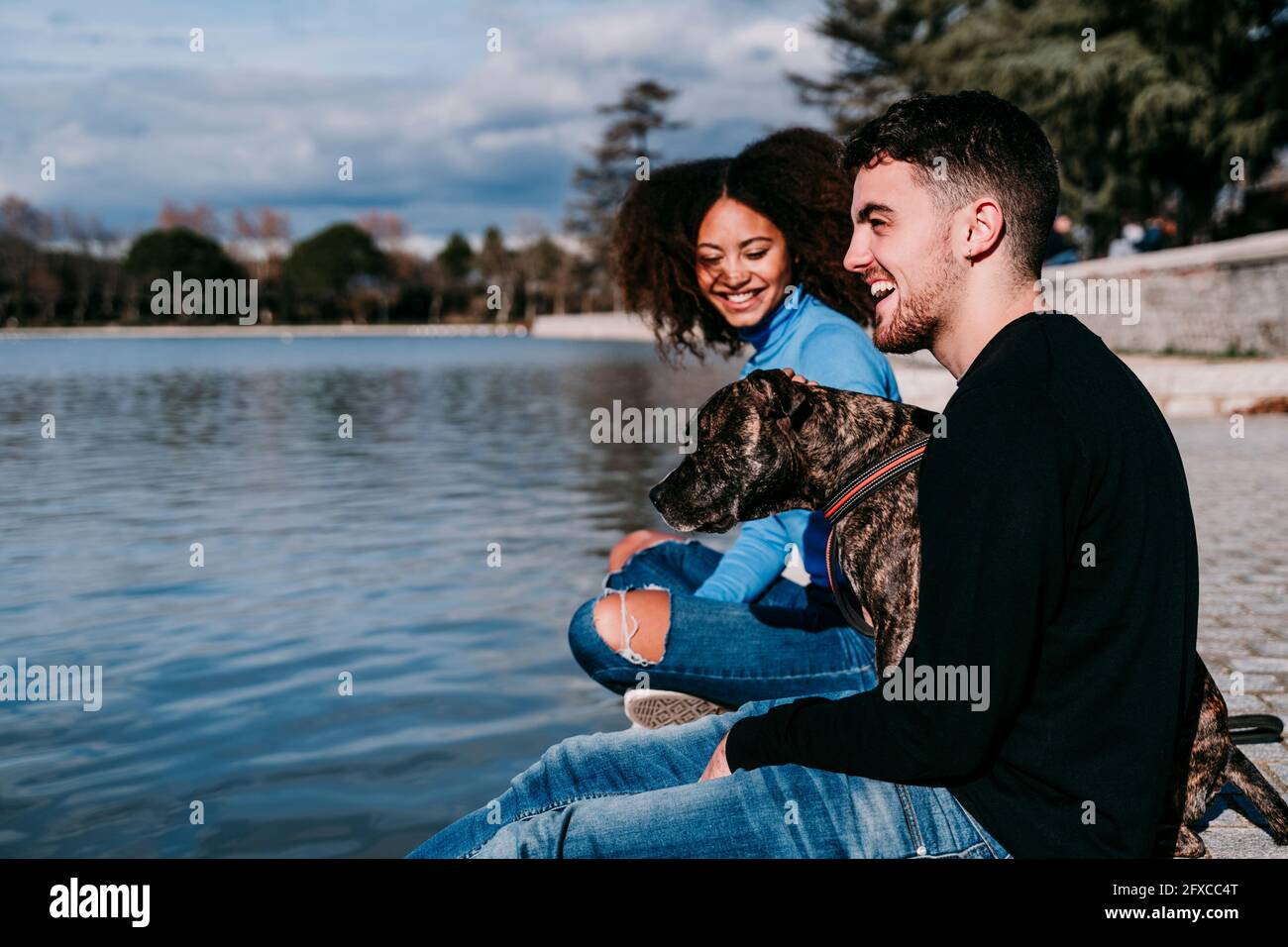 Lächelndes junges Paar, das am sonnigen Tag mit Hund am See sitzt Stockfoto