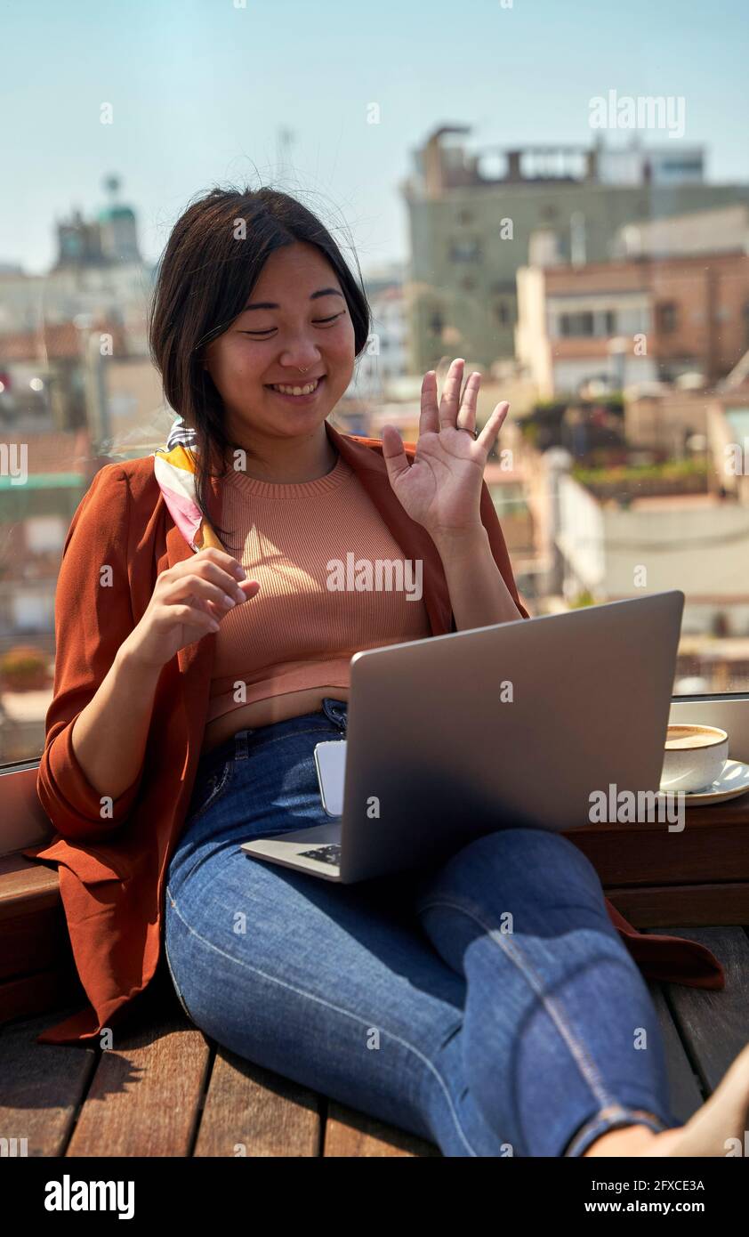 Lächelnde Frau, die Hand zum Videoanruf auf dem Laptop winkt, während sie auf der Dachterrasse sitzt Stockfoto