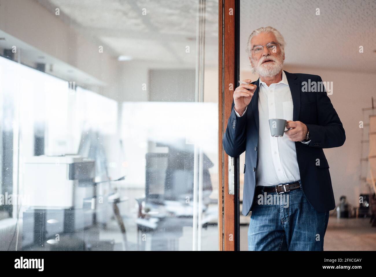 Ein älterer Geschäftsmann hält eine Zigarette mit einer Kaffeetasse, während er sich an einer Glaswand im Büro lehnt Stockfoto