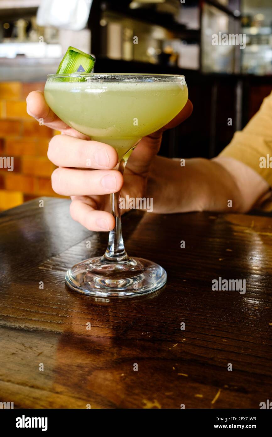 Mann, der einen Gurkenkimlet in einem stämmigen Glas hält, sitzt An einem Holztisch Stockfoto
