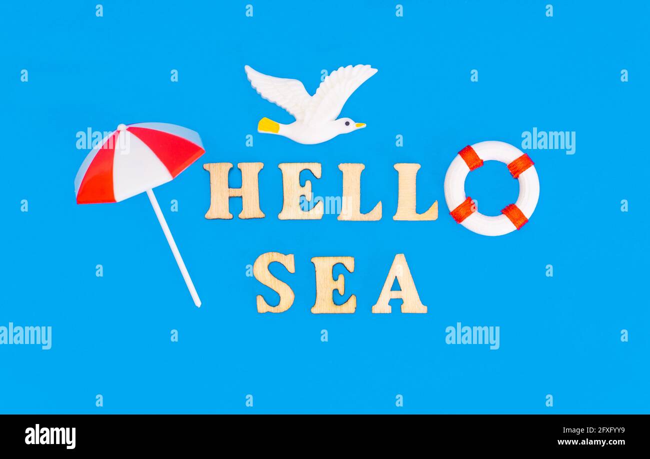 Kreative Say HELLO to the SEA Komposition aus Holzbuchstaben, Spielzeugmöwe, Rettungsring und einem Sonnenschirm auf blauem Hintergrund, der Wasser imitiert Stockfoto