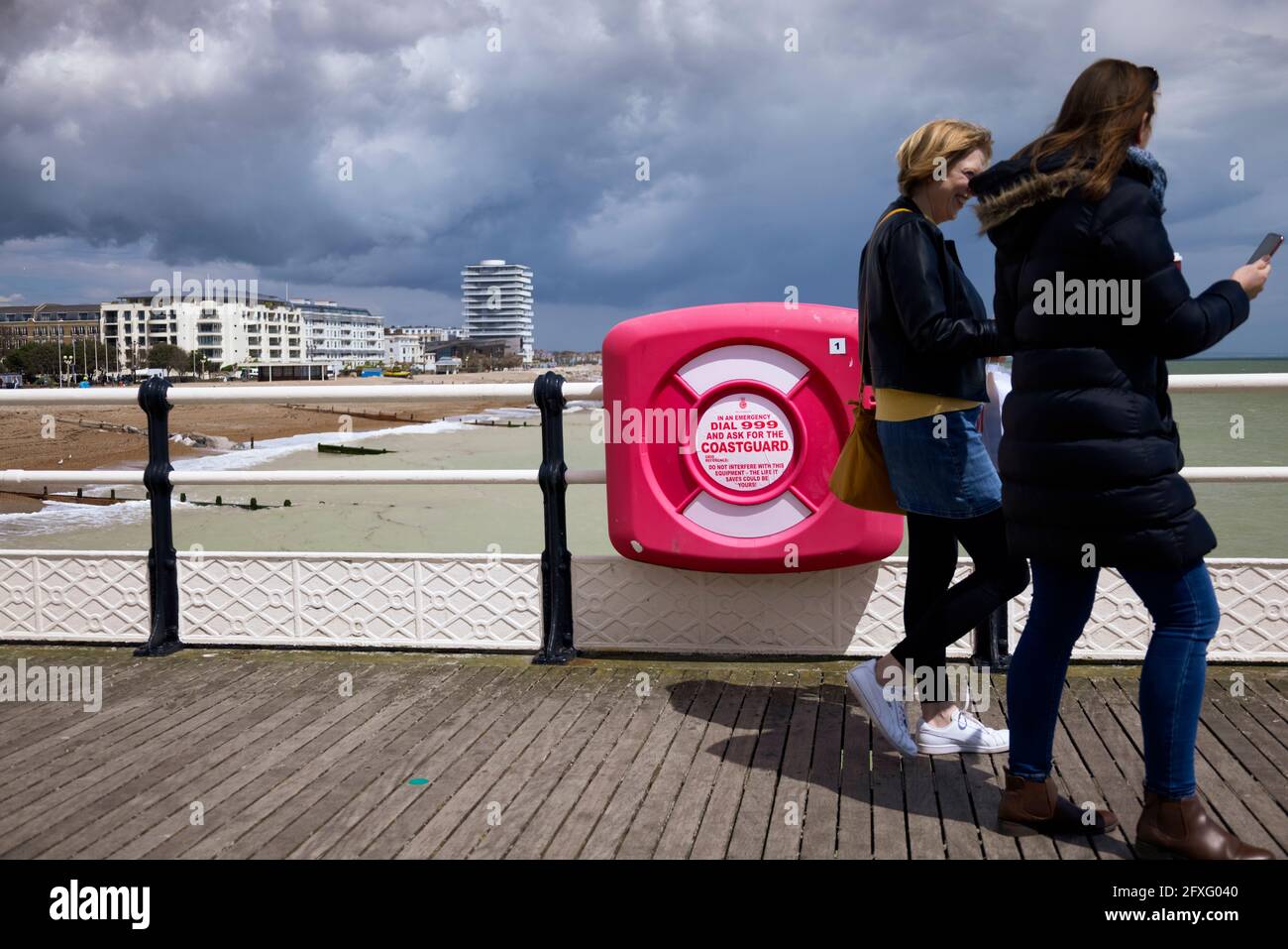 Da sich Gewitterwolken sammeln und eine Schwimmerin in der Nähe von Schwierigkeiten berichtet, gehen zwei Frauen ohne einen Blick an einem Rettungsring vorbei, mit Blick auf das Mobiltelefon. Stockfoto