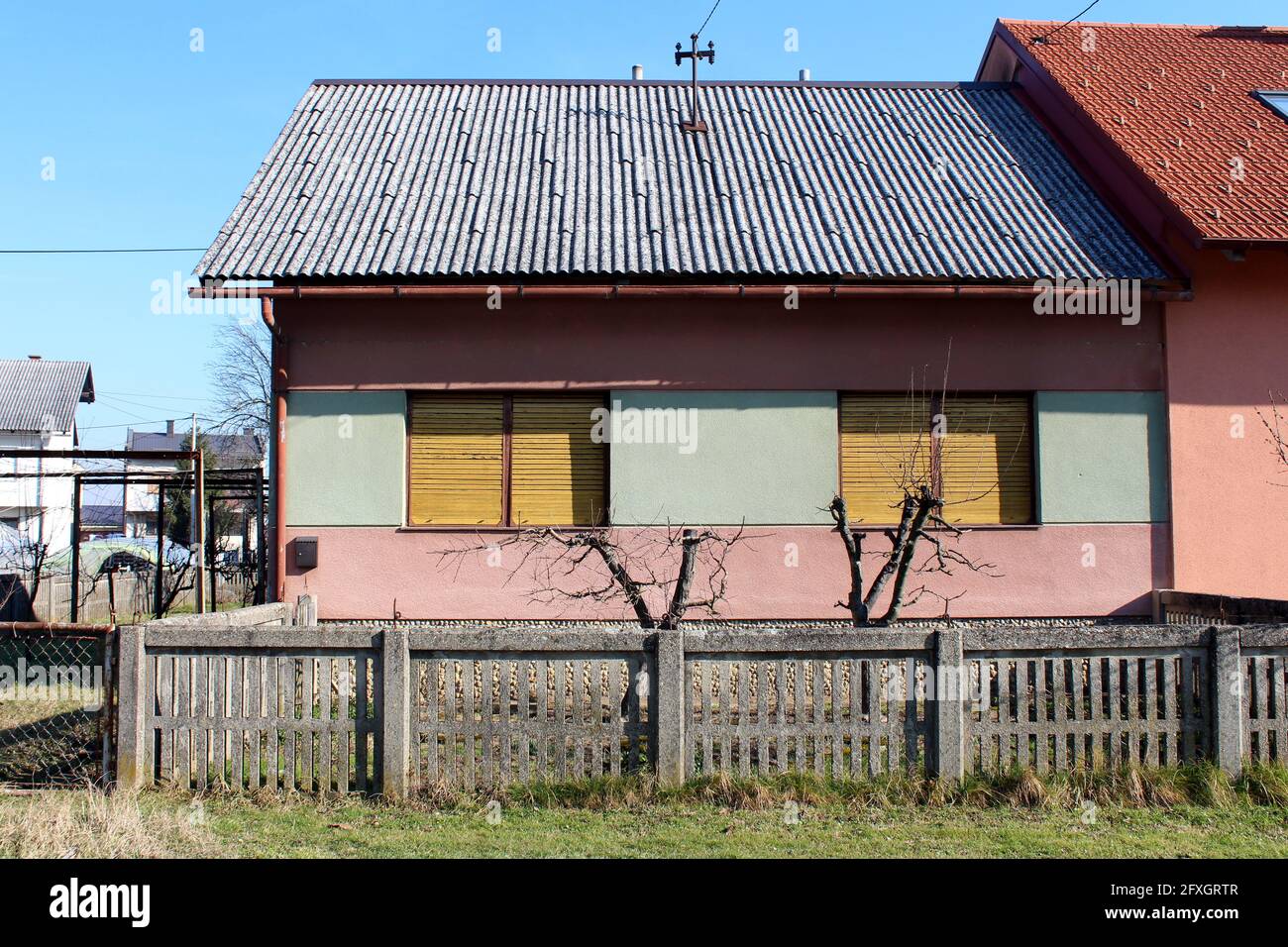 Altes, kleines, an einen Vorort angeschlossenes Familienhaus, das sanierungsbedürftig ist, mit verblassener Fassade und geschlossenen, verfallenen Fensterläden, umgeben von einem Betonzaun Stockfoto