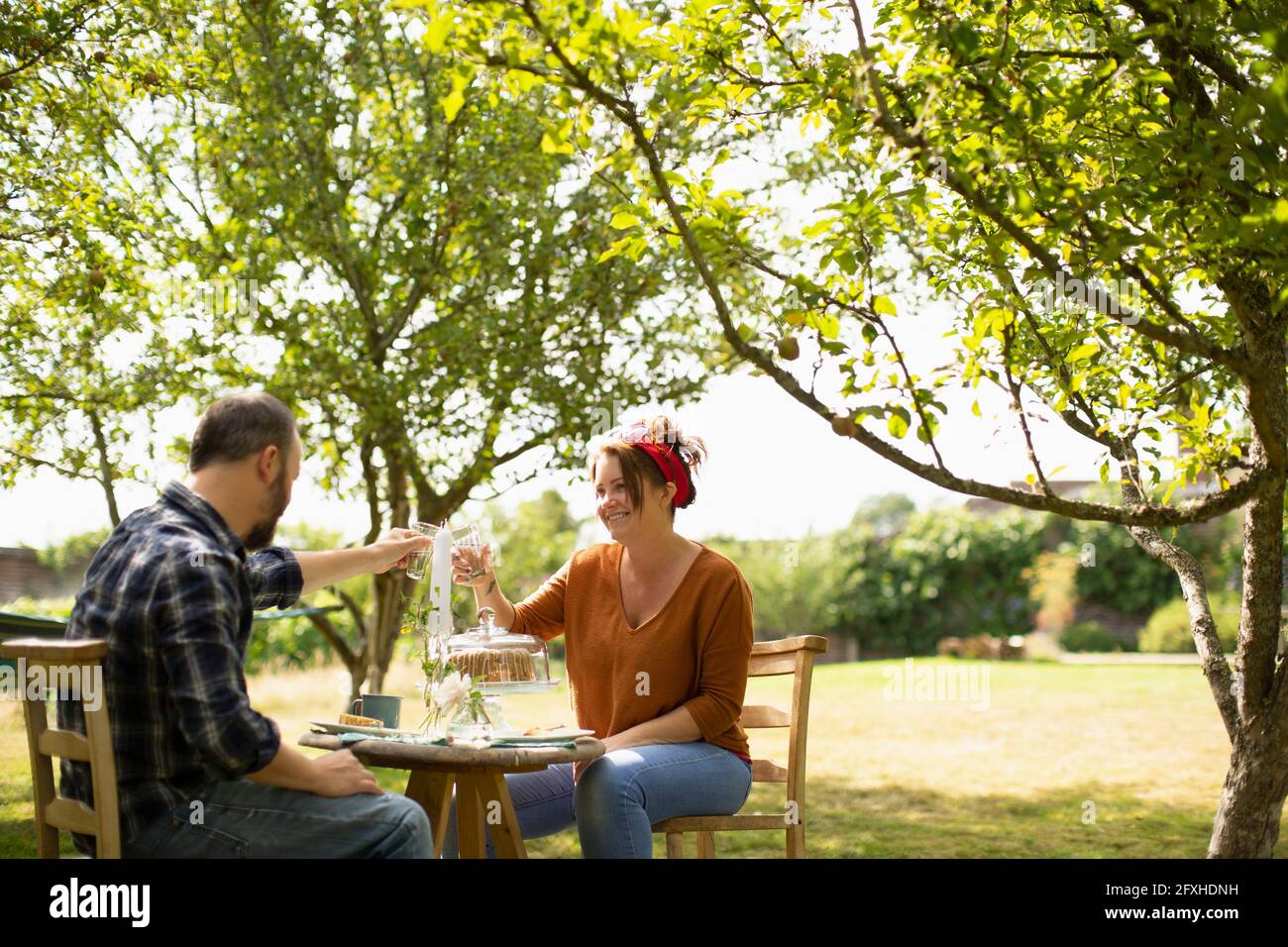 Glückliches Paar, das im sonnigen Sommergarten Wassergläser am Tisch toaste Stockfoto