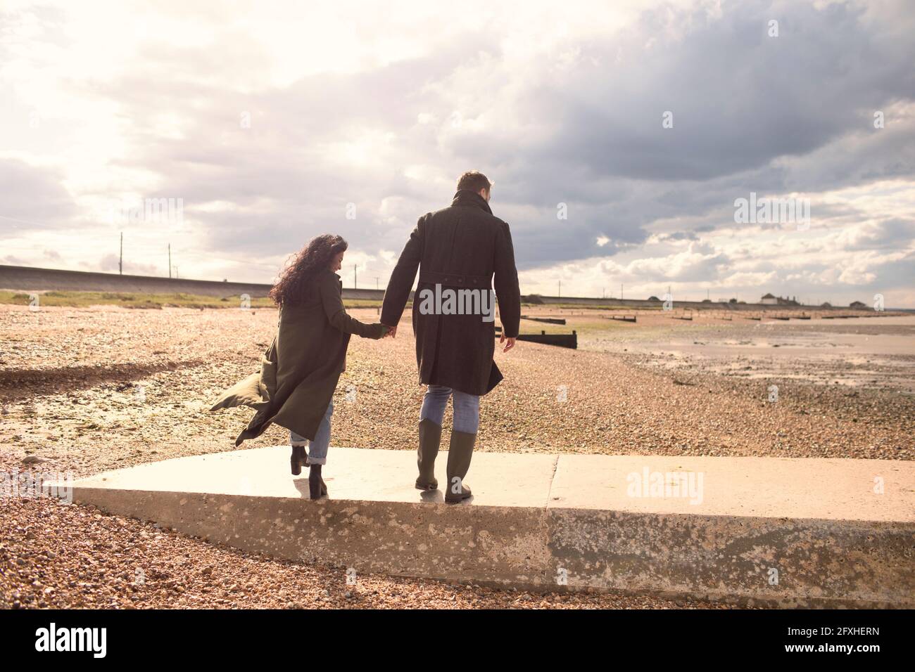 Paar in Wintermänteln, die Hände halten und am sonnigen Strand spazieren gehen Stockfoto