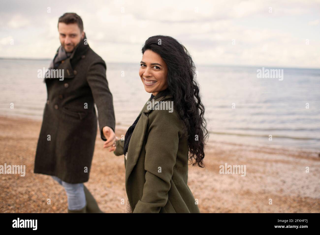 Portrait glückliches Paar in Wintermänteln, das die Hände am Strand hält Stockfoto